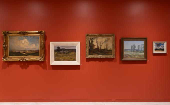 Five landscape paintings are hung against a vermillion wall. The frames are shifted up and down on the wall in order to align the horizon lines of each different bucolic setting. 