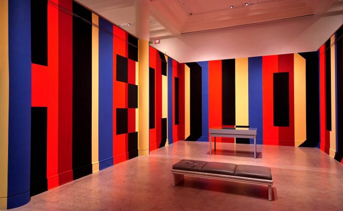  Thick vertical stripes of orange, red, yellow and blue are painted on the walls of the gallery from floor to ceiling. Black paint is used as the shadows of the Superstar Shadow text. We can read the partial words: HE COLOU, wrapping the bend.