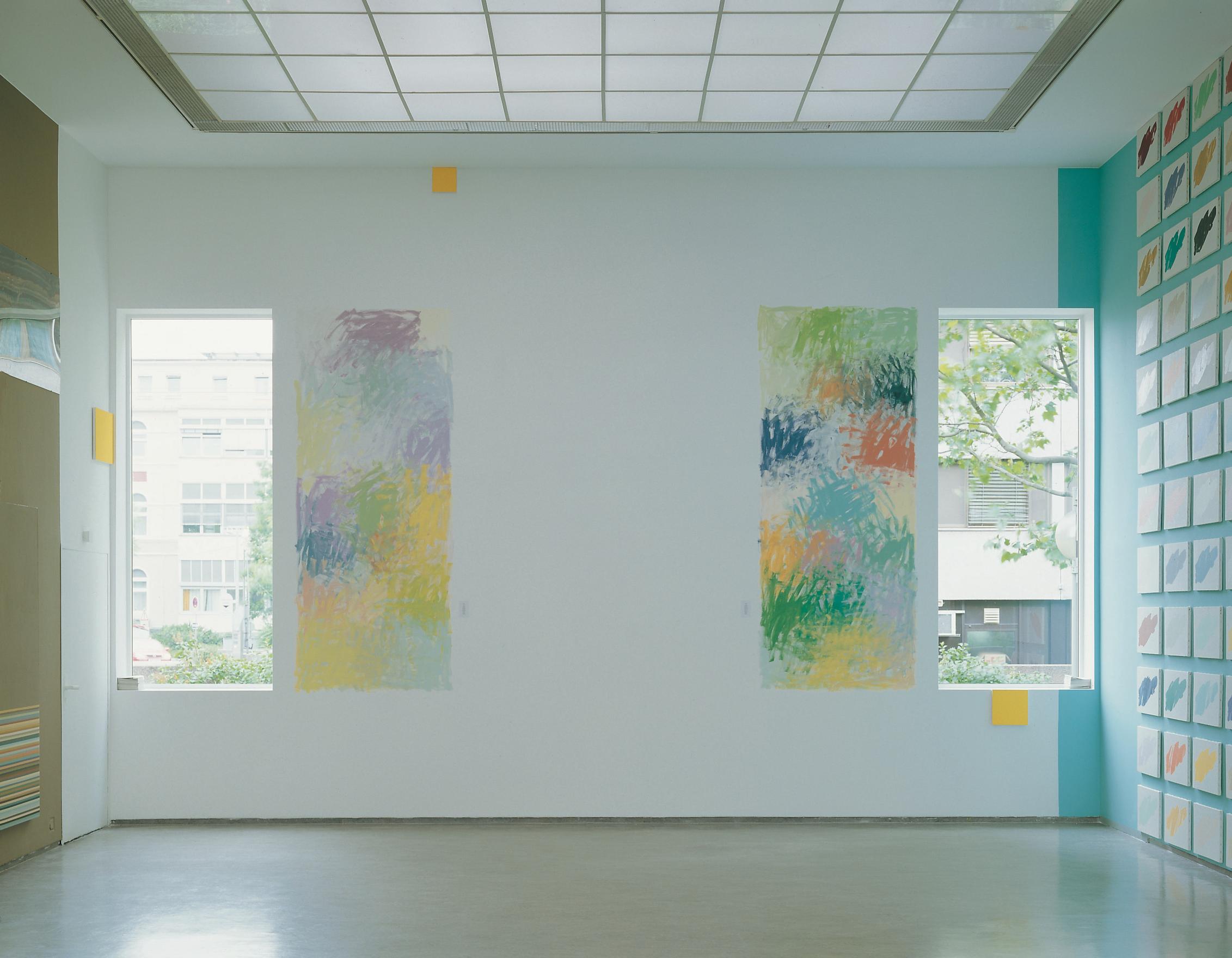 A square gallery is lit by the sunlight streaming in through two rectangular windows in the back wall. Beside each window is an expressive wall painting with the same dimensions, made of layers of overlapping scribbles each with its own distinct colour pallet; the left: lighter pastel tones of eggshell blue, canary yellow and leaf green, the right: rust oranges, deep ocean blues and evergreens. There are three yellow squares positioned at various heights around the room and the adjacent walls hold otherwork