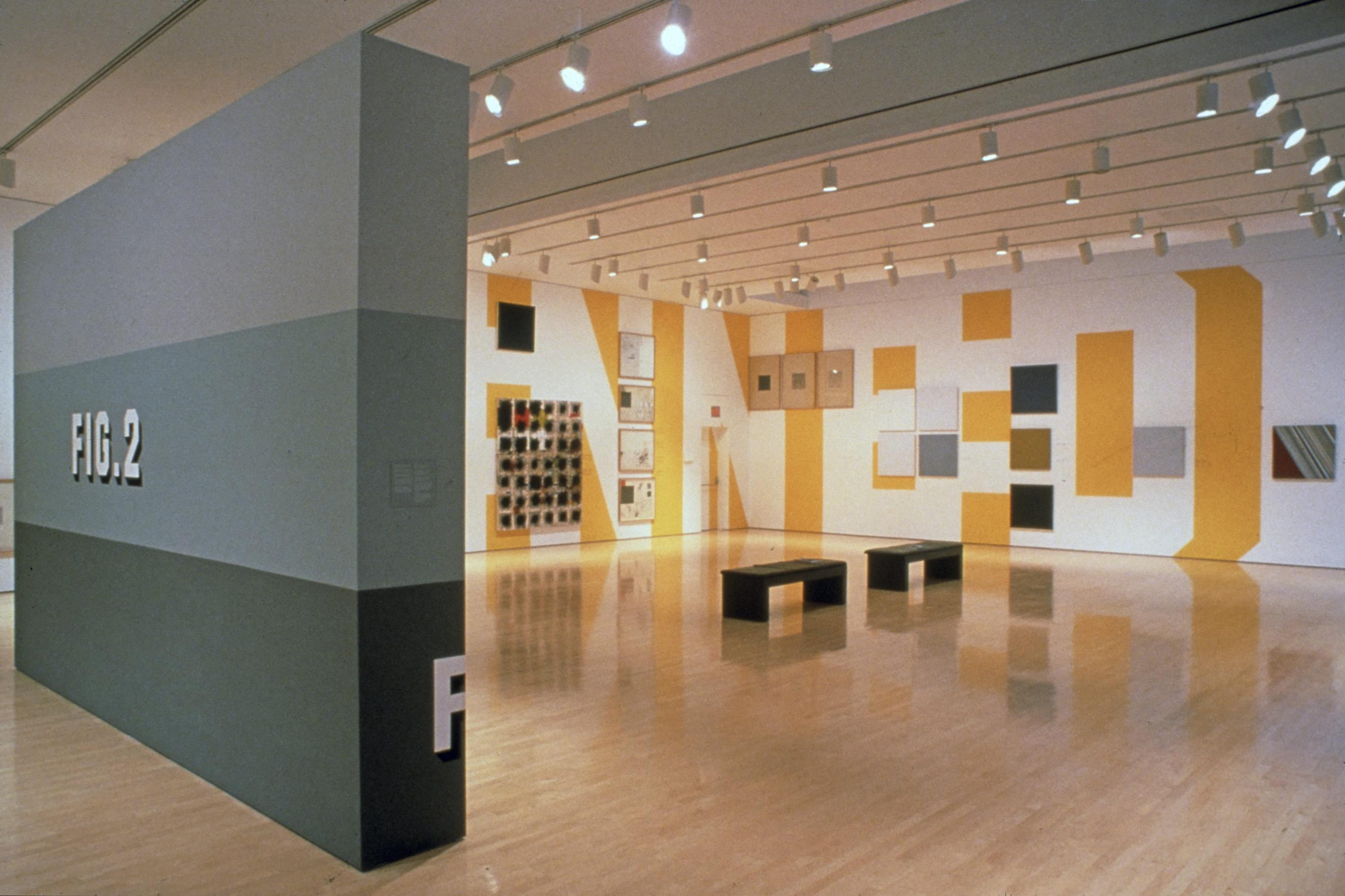A rectangular wall with three grey stripes, painted dark to light moving up, with the word: FIG.2 painted in the middle stripe, towers in the left corner of a gallery room. Behind the wall, two walls are lined with artworks of various sizes. Hard to decipher at the distance, the works tone in black, red and greys against the wall with floor to ceiling letters painted with yellow and white: N N E D.