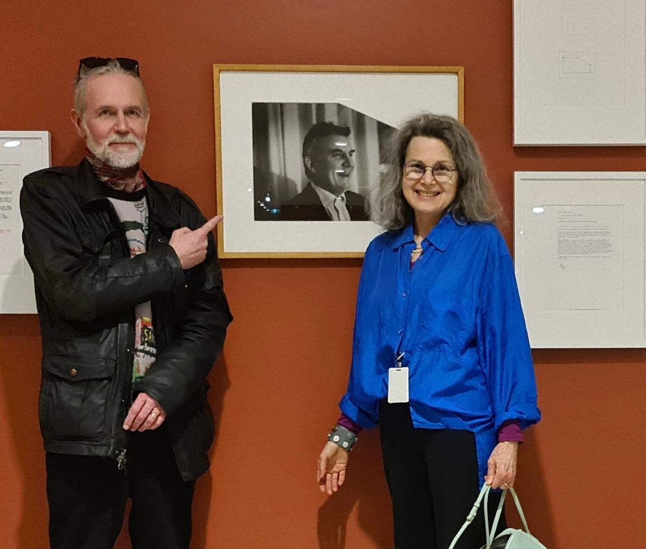 Erling Klingenberg and Cathy Busby stand in front of a black and white portrait of Garry Neill Kennedy. Klingberg is wearing a black leather coat and has black sunglasses resting on his grey hair. Busby is wearing a royal blue silk top and wears black wire rimmed glasses and a broad smile. 