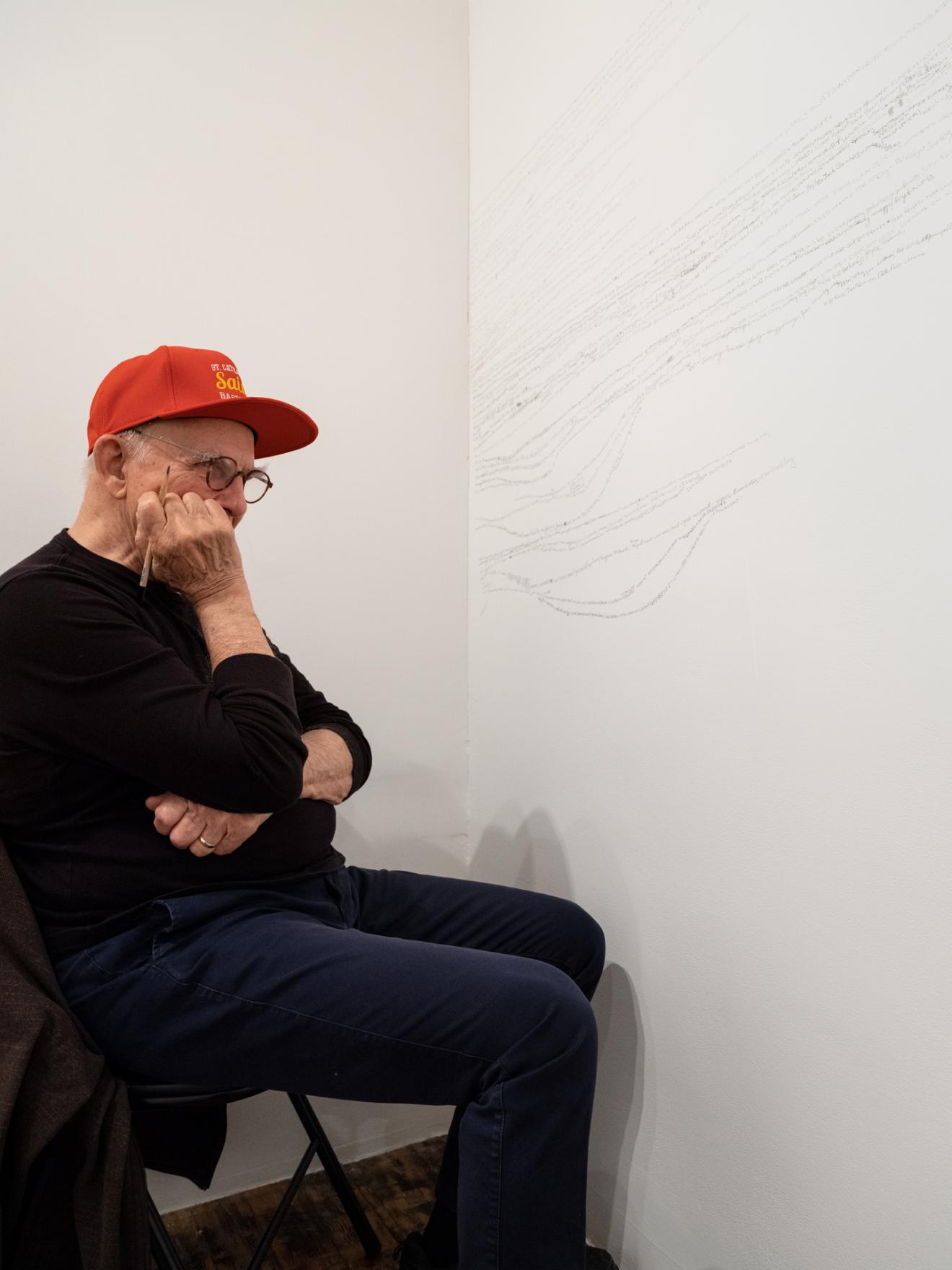  Garry, an older white man wearing glasses,  a red Saint Catharine’s Saints baseball cap and black sweater, sits in the corner of a white walled room. He rests his chin on his hand which holds a pencil as he stares at the wall of undulating lines of people's names.