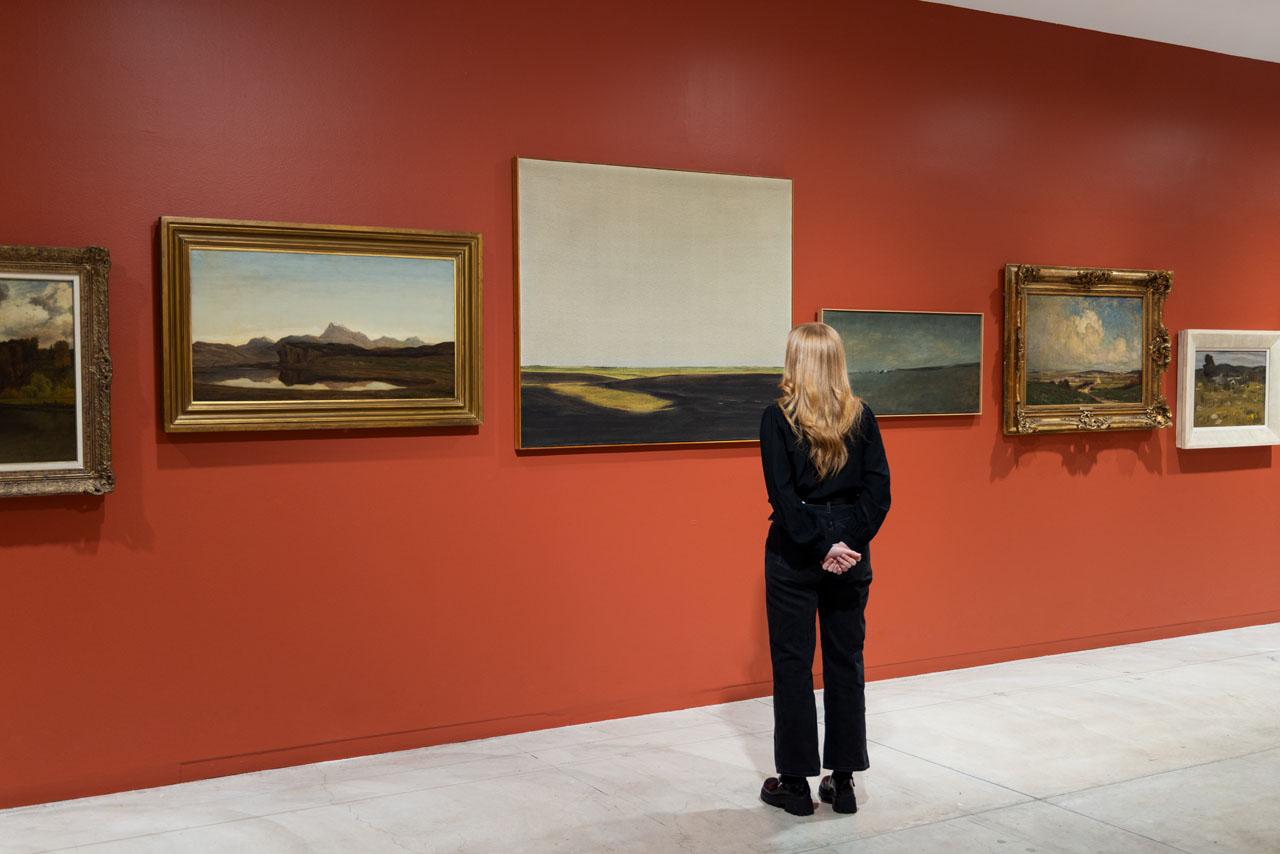 A viewer stands in front of a large landscape painting with a vast grey sky and dark green rolling grasses. The surrounding paintings are shifted up and down on the wall in order to align the horizon lines of each neighbouring painting. 