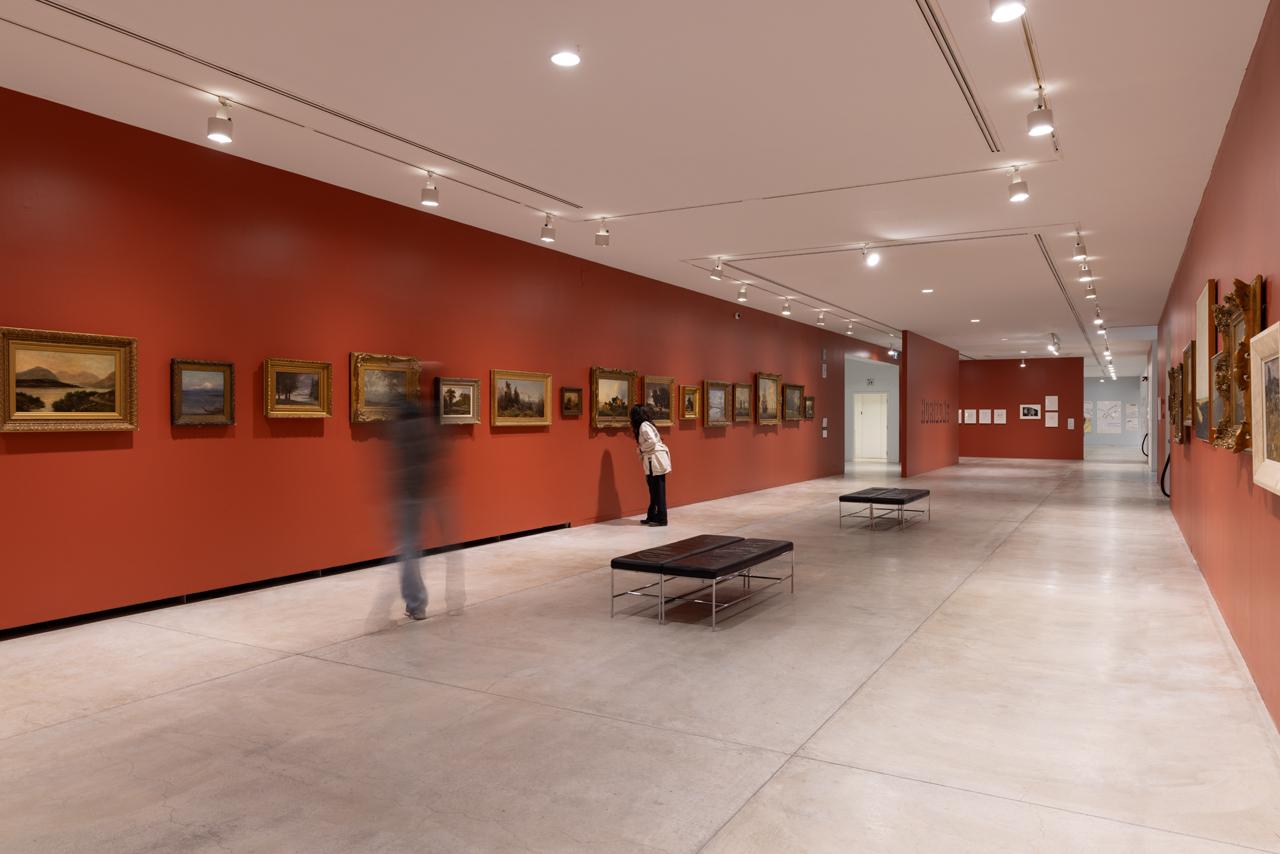 Landscape paintings line the vermillion walls of a long gallery space. The paintings are framed with antique filigree frames and hung at various heights to ensure the horizon lines of each landscape line up with those in the neighbouring paintings. A few people move about the space or stop to look.