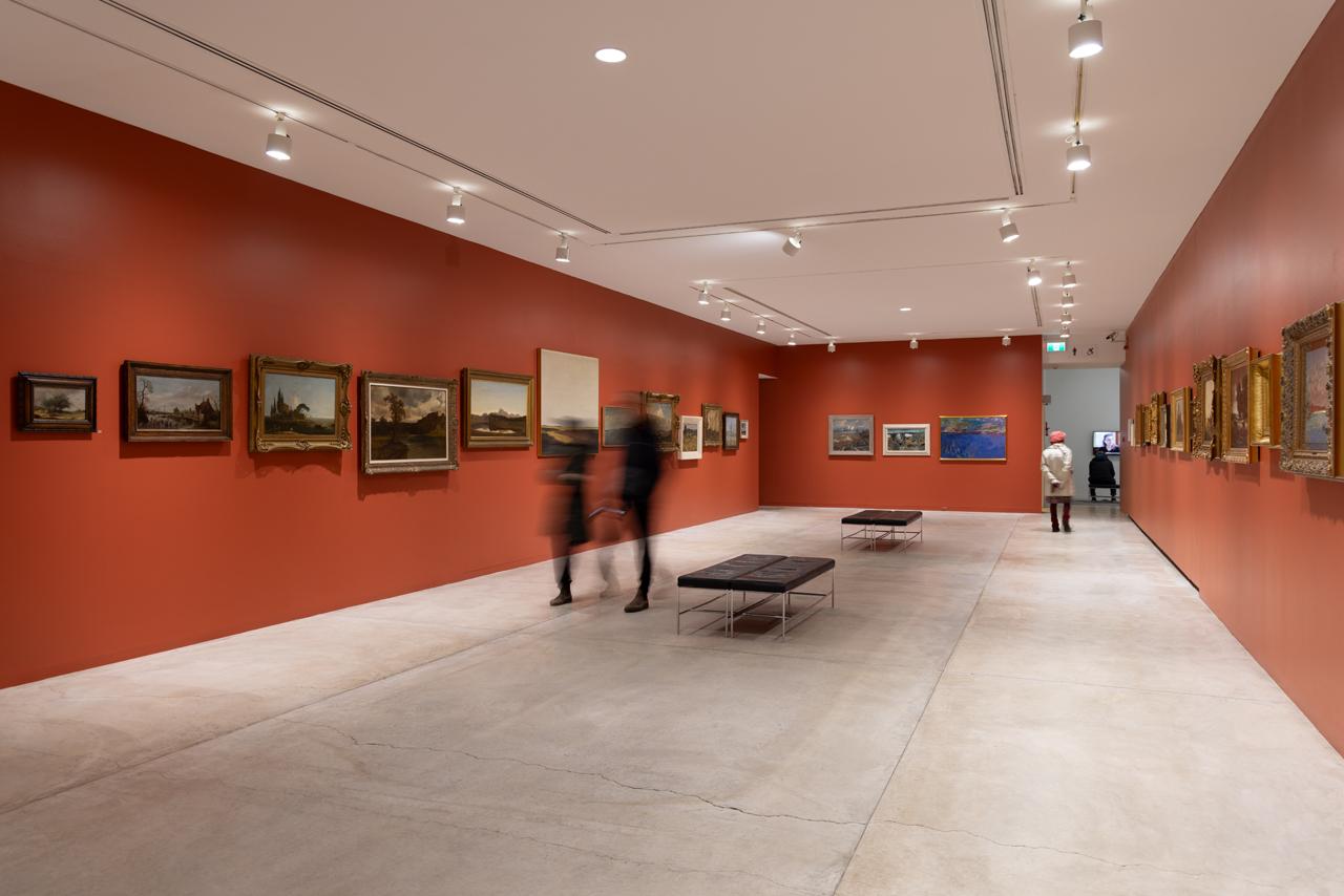Landscape paintings line the vermillion walls of a long gallery space. The paintings are framed with antique filigree frames and hung at various heights to ensure the horizon lines of each landscape line up with those in the neighbouring paintings. A few people move about the space quickly blurring their bodies. 