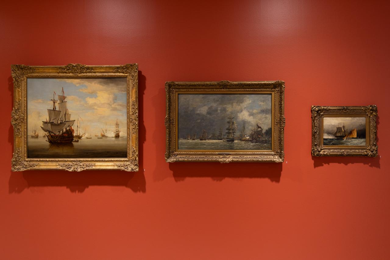 Three ocean landscape paintings are hung against a vermillion wall. Each has a golden filigree frame which decreases in size from left to right. The horizon lines of each different sea and sky align although the composition of ships and stormy or calm waters ranges. 