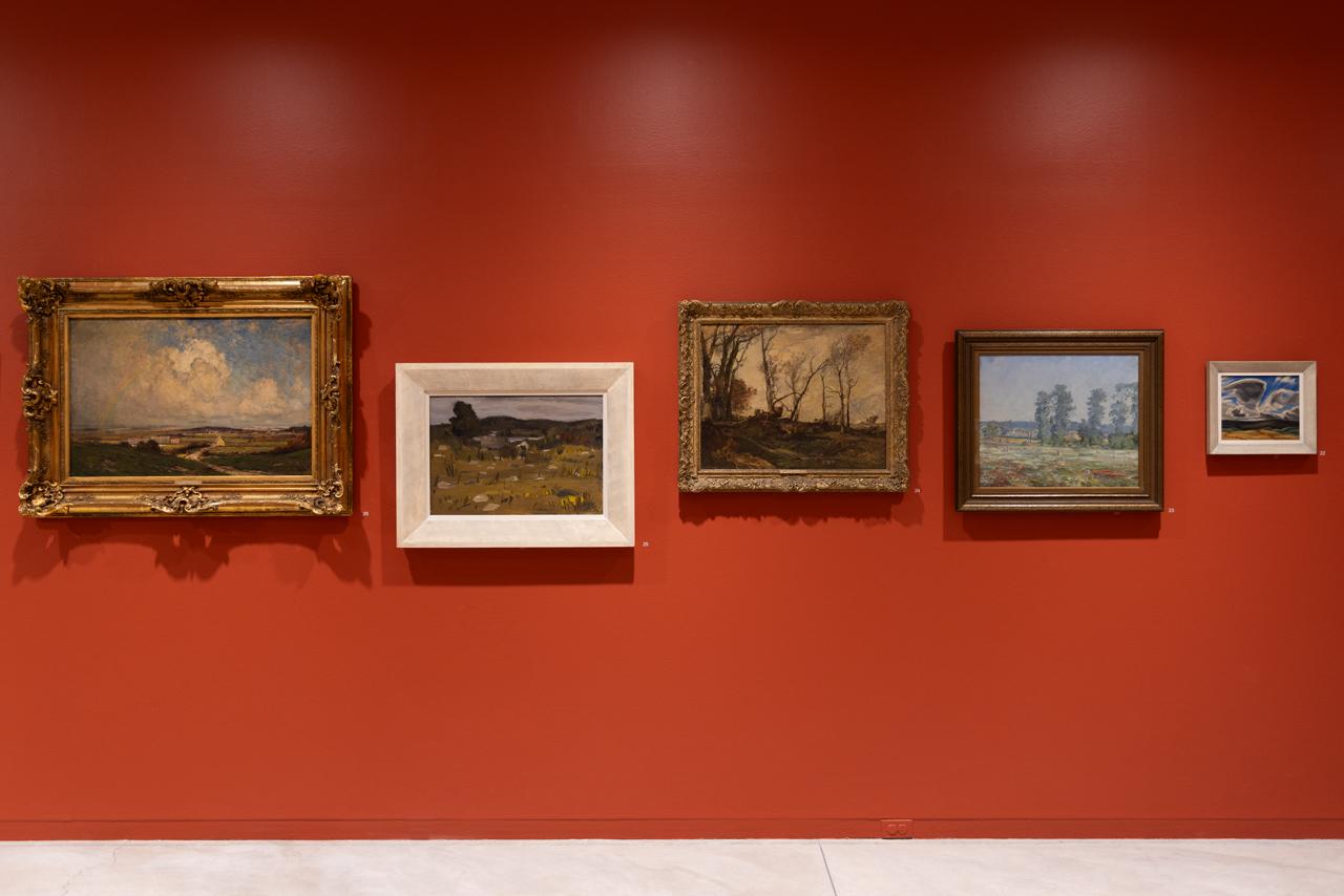Five landscape paintings are hung against a vermillion wall. The frames are shifted up and down on the wall in order to align the horizon lines of each different bucolic setting. 