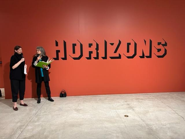 Cathy Busby and Mandy Ginson, two women wearing black and both holding microphones, stand next to the black lettering of the title vinyl of the Horizons exhibition. 