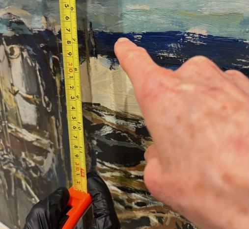 Close up, a finger points to the thin line where deep ocean and light sky meet in a painting. A tape measure runs vertically on the left edge measuring the distance from the floor to the horizon line during installation.