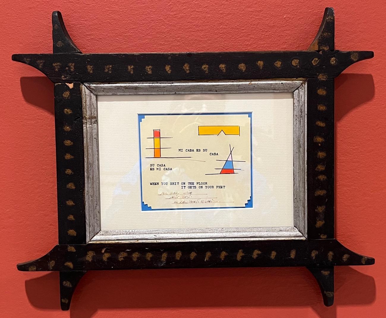 A small note paper framed with blue border holds orange and yellow shapes and the words quote: su casa es mi casa, mi casa es su casa, when you shit on the floor it gets on your feet, are written using a typewriter. The whole note is framed with black wood that has swooping pointed corners which look hand carved.