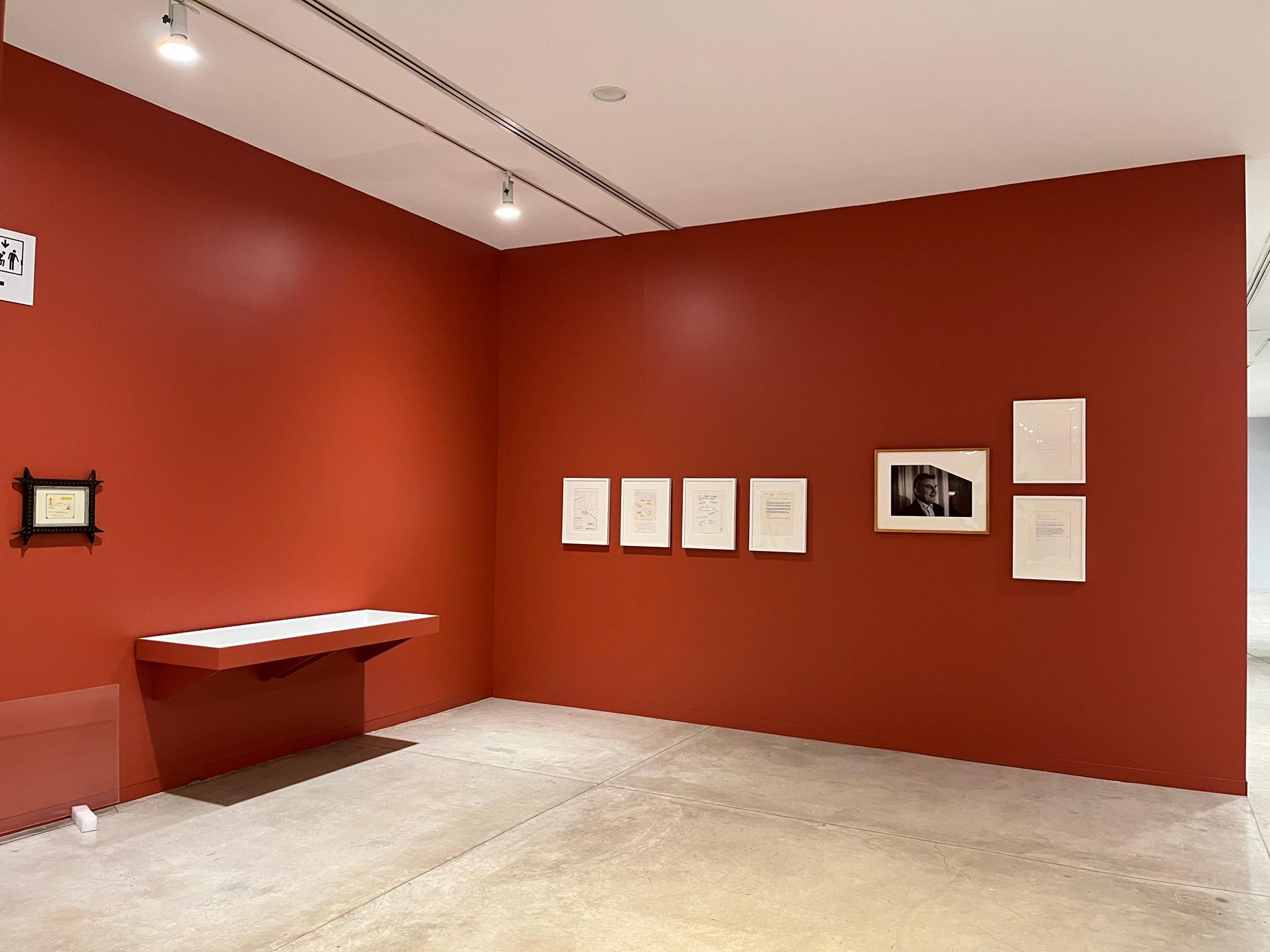Archival documents hang on vermillion walls in an L-shaped gallery room. The documents are at a distance which obscures their details. 