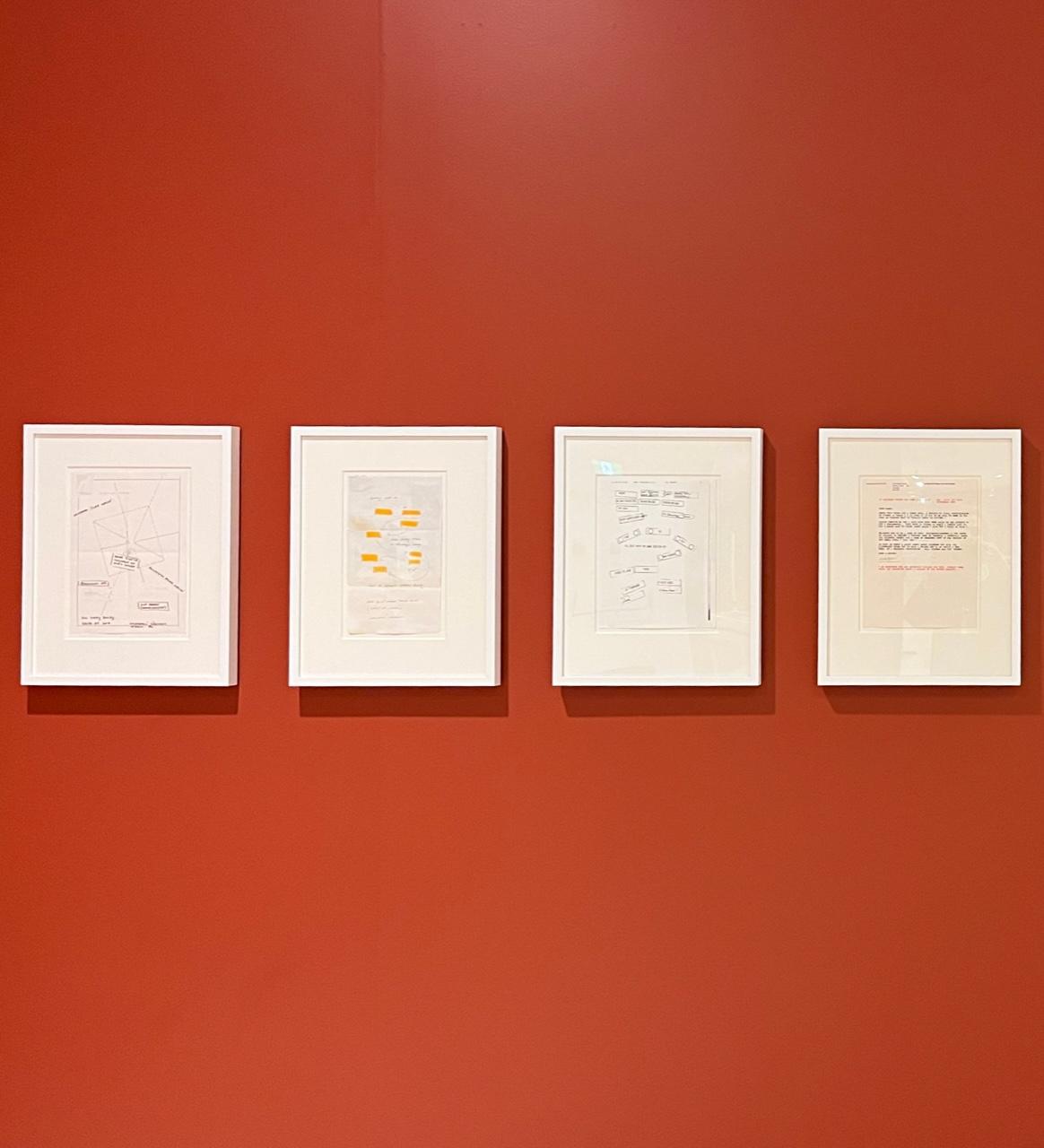 Four framed documents ranging from note paper drawings to typed letters hang in a row on a vermillion wall. 