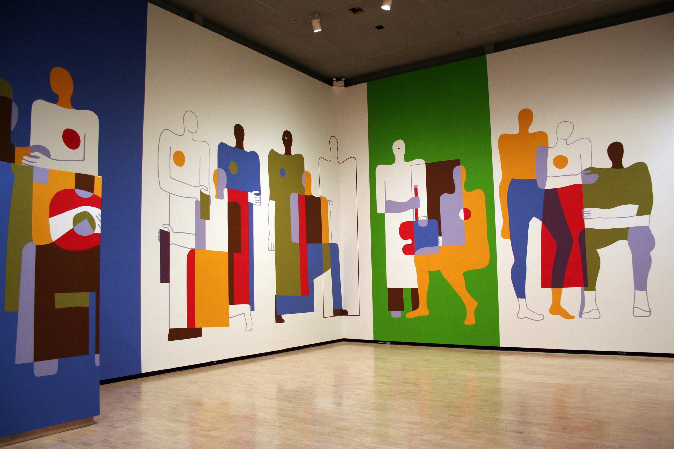  The walls of a large open gallery space are filled from floor to ceiling with a colourful mural. Blue, white and green background sections frame abstract figures supporting one another. Their multicoloured bodies overlap, their blocks of colour changing upon contact.