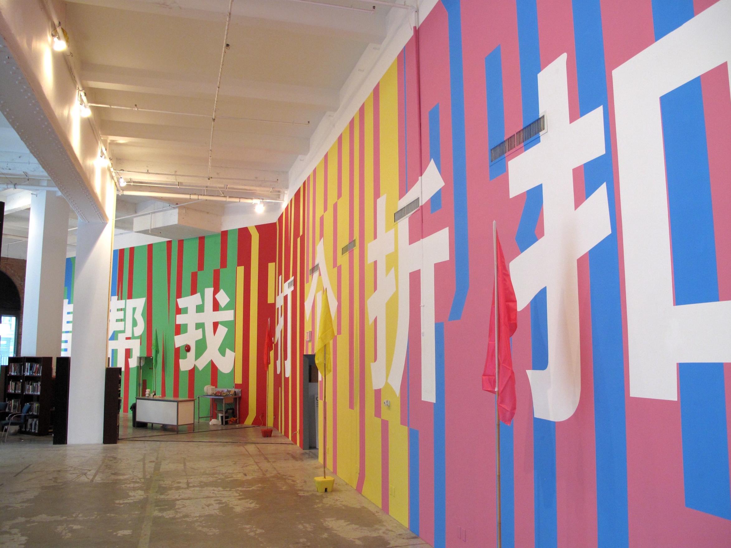 A colourful wall filled with text stretching from floor to ceiling in both English and Mandarin stretches around the right side of the gallery space. The English text is in Superstar shadow font and coloured in green, red, yellow, pink and blue. The Mandarin characters are in white over top. Small flags on bamboo poles stand near the wall, matching the colours of the paint. 