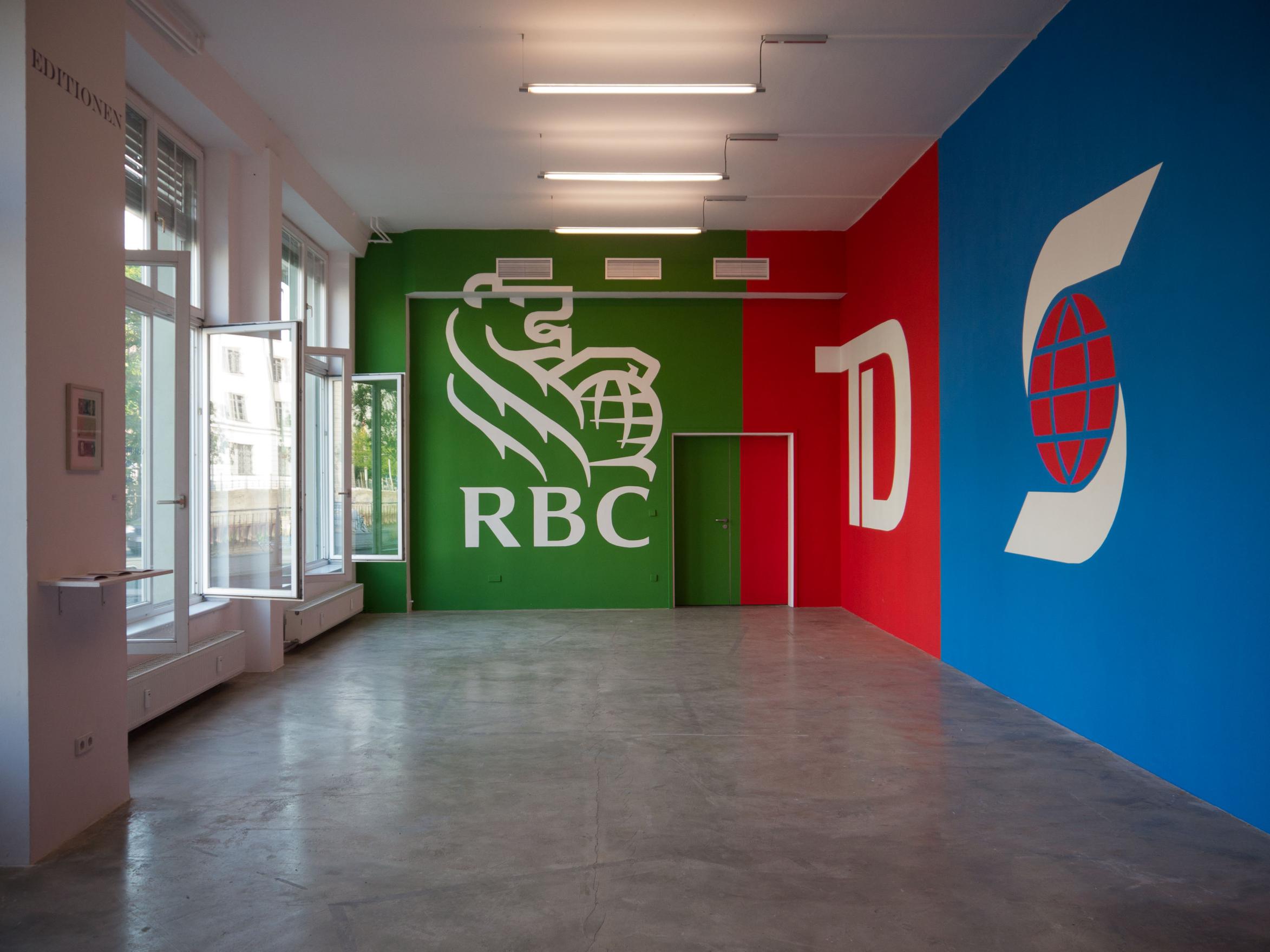 The walls of a concrete floored gallery space are painted with the logos of three top Canadian banks, but the colours have been swapped. The lion and globe of the RBC logo is in white and green, TD bank stands out in white text on bold red, and Scotiabank's ‘S’ globe is in white and red on a blue background. 