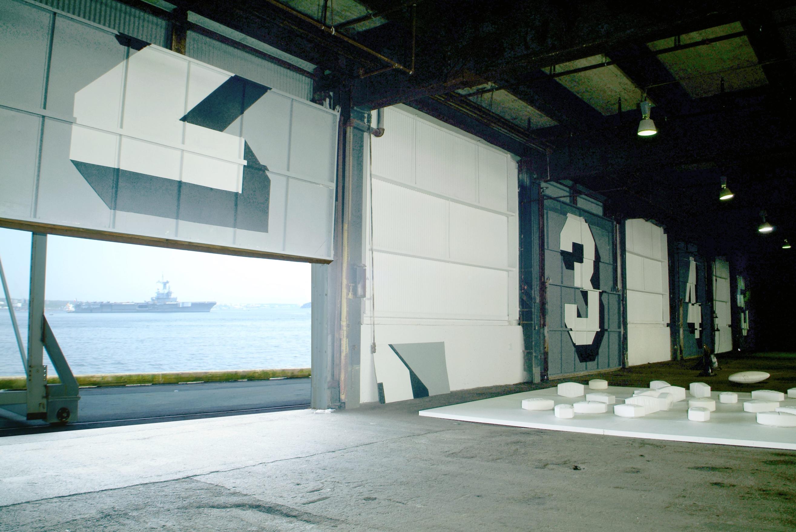 A loading bay door with a large number 2 painted on it rolls up to reveal the ocean and port beyond. Three more loading doors with similarly large numbers stretch to the back of the warehouse space, they are numbered 3, 4, 5 in white and black Superstar Shadow font.