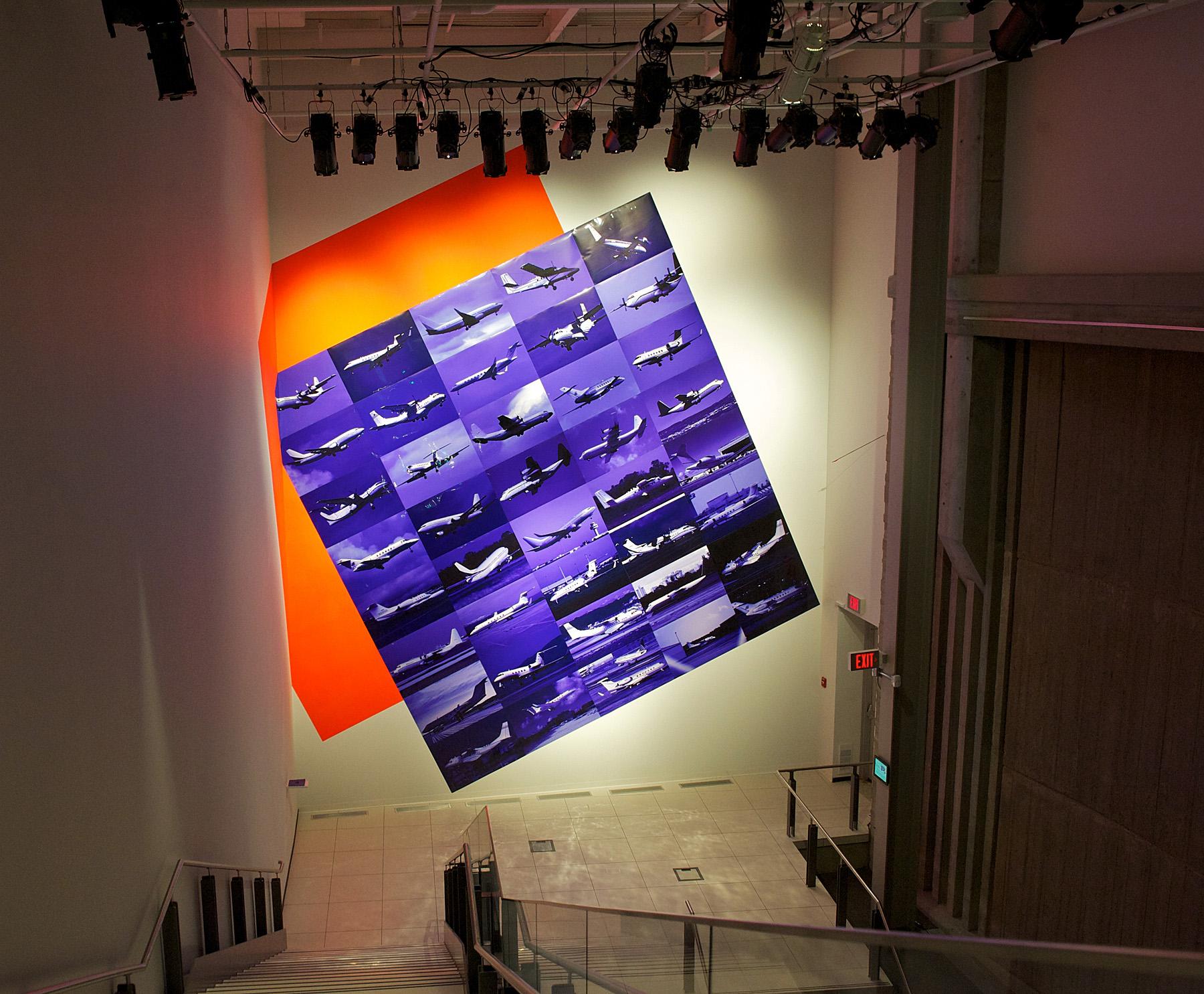 A grid of 45 photographs of airplanes taking off and landing tilt and wrap around the high-ceilinged wall of an open stairwell. A large orange square acts like a shadow to the grid, making the blue pictures pop. 