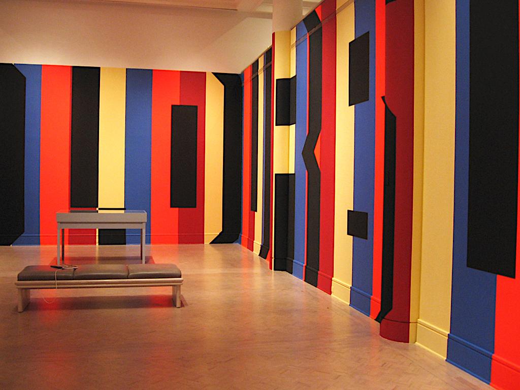  Thick vertical stripes of orange, red, yellow and blue are painted on the walls of the gallery from floor to ceiling. Black paint is used as the shadows of the Superstar Shadow text. In the corner shown the OUR at the end of the word colour can be seen wrapping the bend.