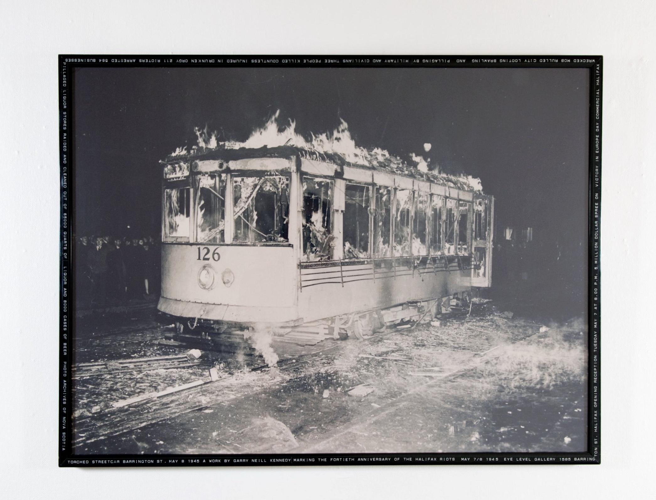  A large black and white photograph of a street car engulfed with flames is framed by a thin black strip of Dymo-tape flecked with tiny white lettering, illegible from this distance. 