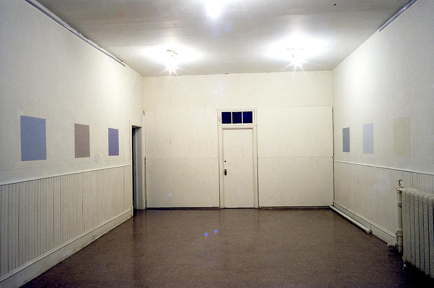 A small white walled gallery is encircled with squares painted on the walls at eye level for a person around 5 foot 10 inches. The squares are all muted tones of blue, beige, and shades of white. 