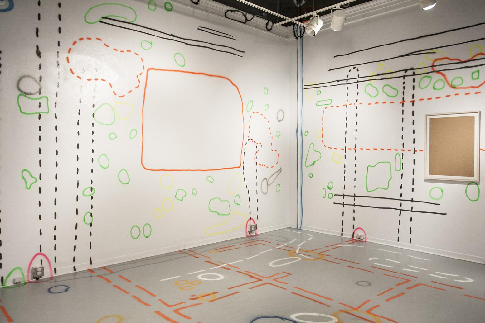 A gallery is spray painted like a construction road site. There are dotted lines, and globular shapes in green, orange, pink, white, black, blue and yellow which denote flaws in the gallery space, from electrical units to exposed pipes to dents and cracks in the walls. There is a framed piece of cardboard on the right wall. 