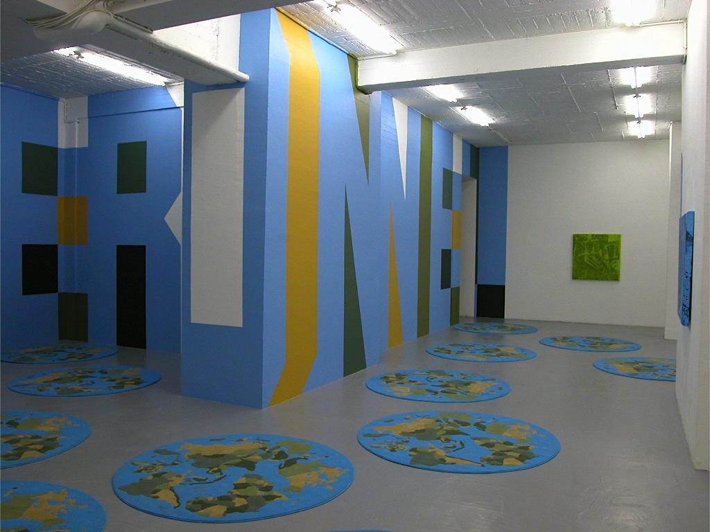 The tail end of the phrase REIN DE PERSONNEL, painted in large Superstar Shadow lettering, can be seen snaking over the walls of a gallery space. The letters are in light blue with drop shadows in green and yellow. Circular world map iKea carpets dot the floor, toning with the colours on the wall.