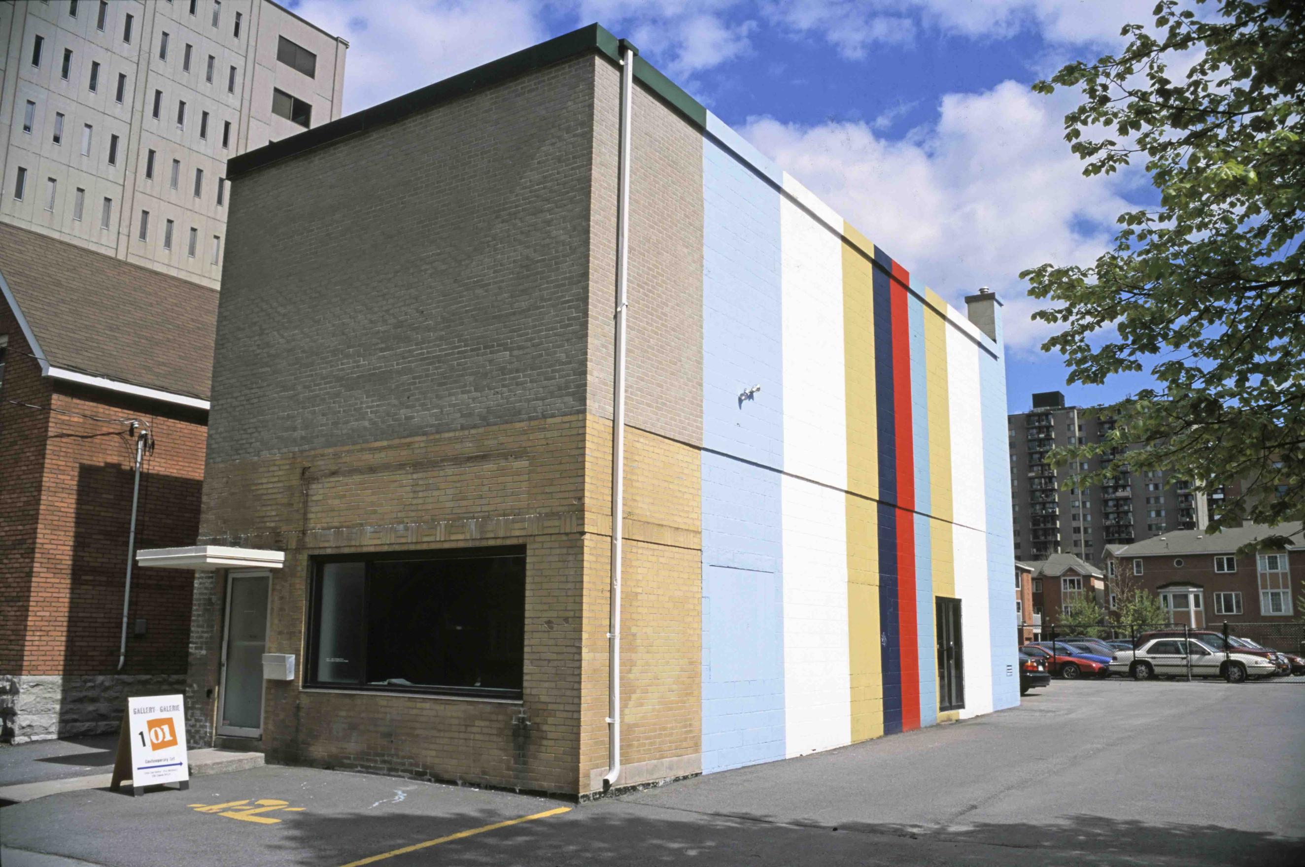 Gallery 101, a solid rectangular brick building, stands at the edge of a parking lot in an urban neighbourhood. The building's bland grey brickface is contrasted by a large stripe painting which fills the west side wall. Bold strips of light blue, white, gold, navy, and red, the colours of the Somalia Medan ribbon, run the vertical length of the building from the concrete lot to the cloudy blue sky above. 