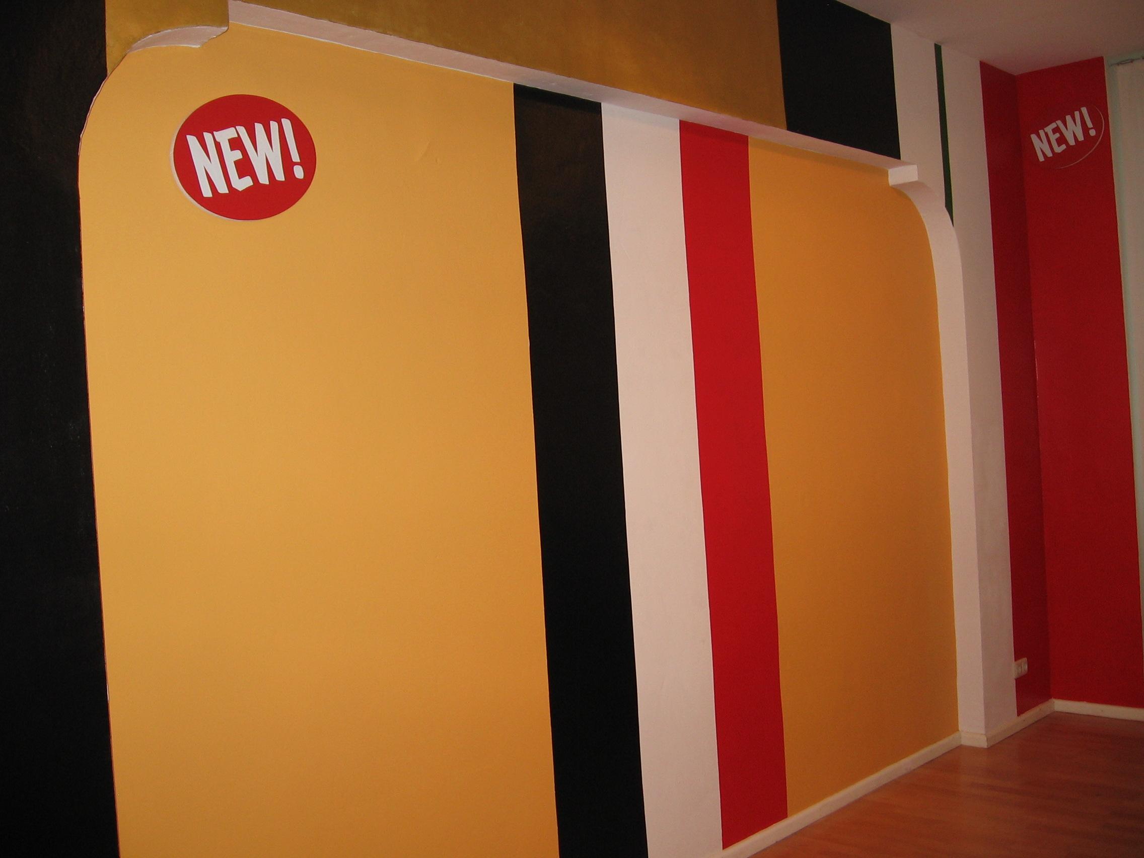Thick black, yellow, white and red vertical stripes fill the gallery wall from floor to ceiling, turning the wall-space into a medal ribbon pattern. Two small red oval canvas’ with NEW! In sign painters lettering are hung on the top of the left wall and on the back wall. 
