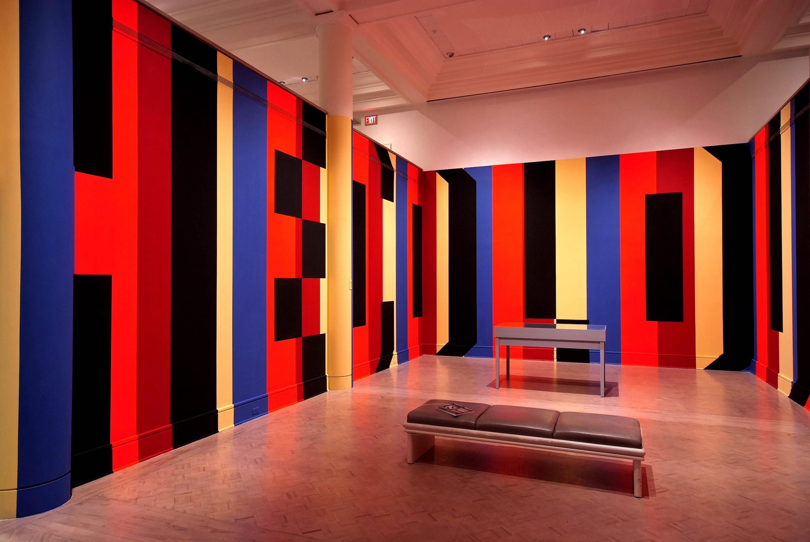  Thick vertical stripes of orange, red, yellow and blue are painted on the walls of the gallery from floor to ceiling. Black paint is used as the shadows of the Superstar Shadow text. We can read the partial words: HE COLOU, wrapping the bend.