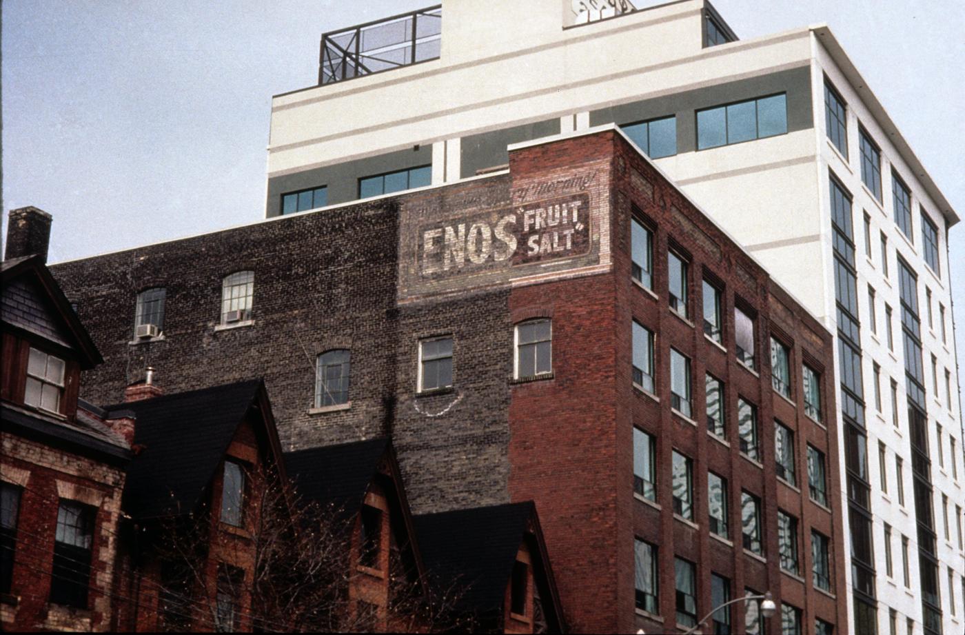 An old brick building is sandwiched between a row of brick townhouses and a newer white and grey apartment building. A faded advertisement painted directly onto the red and brown bricks rests high on the central building's wall. The flacking white paint reads First thing in the morning! ENOS FRUIT SALT.