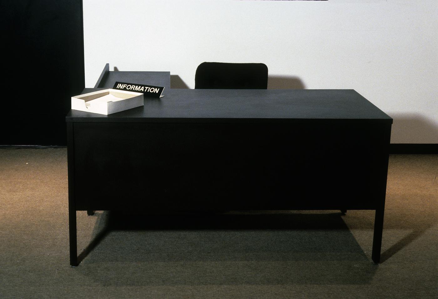 An office desk covered in dark brown formica laminate sits by the entrance of the white walled gallery. Its surface is bare except for a name plate which reads INFORMATION and a stack of papers in a file box. There is a black chair behind the desk that no one sits in. 