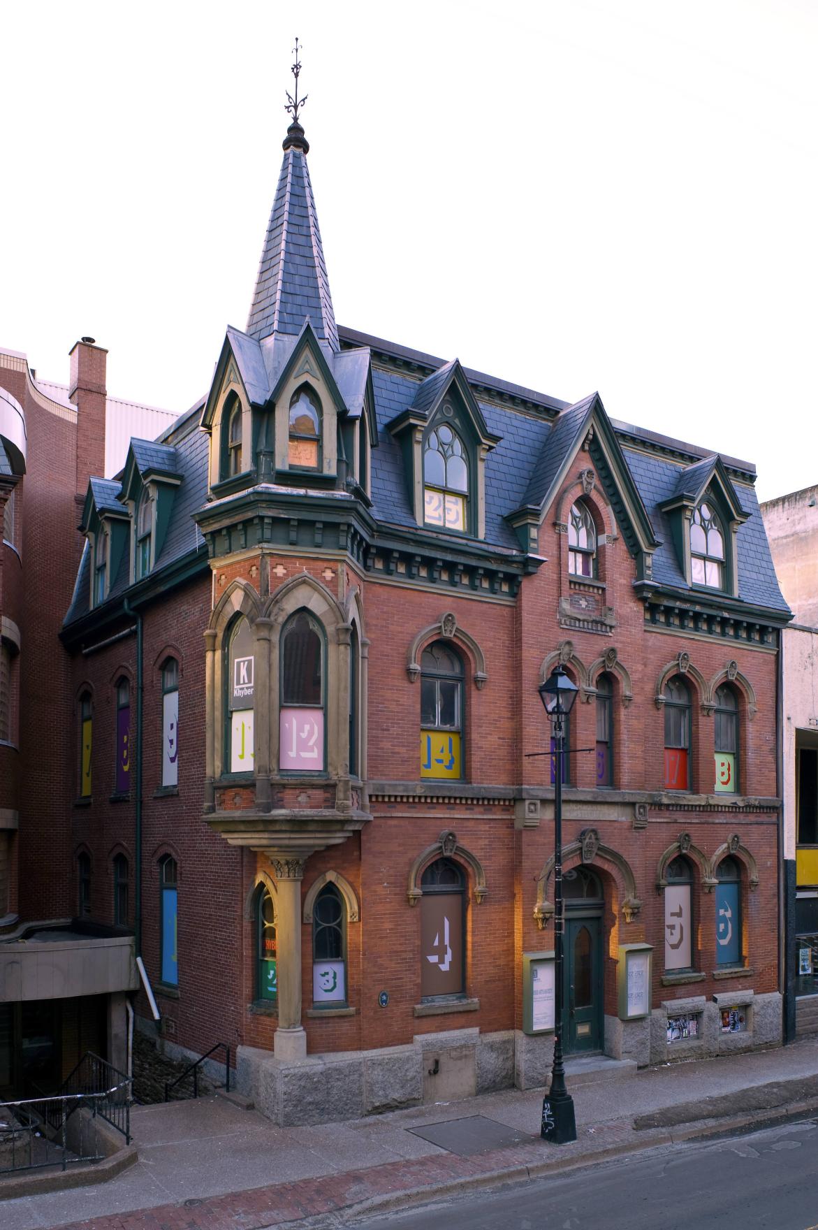 A large brick building with gothic moldings, including a turret with a pointed roof and weather vein, stands majestically in the middle of a Halifax city block. A series of numbered paintings are inset into each of the 27 windows across the building's three floors. Each painting has a large number in Superstar Shadow font in the centre. The numbers and the panel are painted in one colour and the number’s shadow are in another, for example number fourteen is mustard yellow with a forest green shadow.