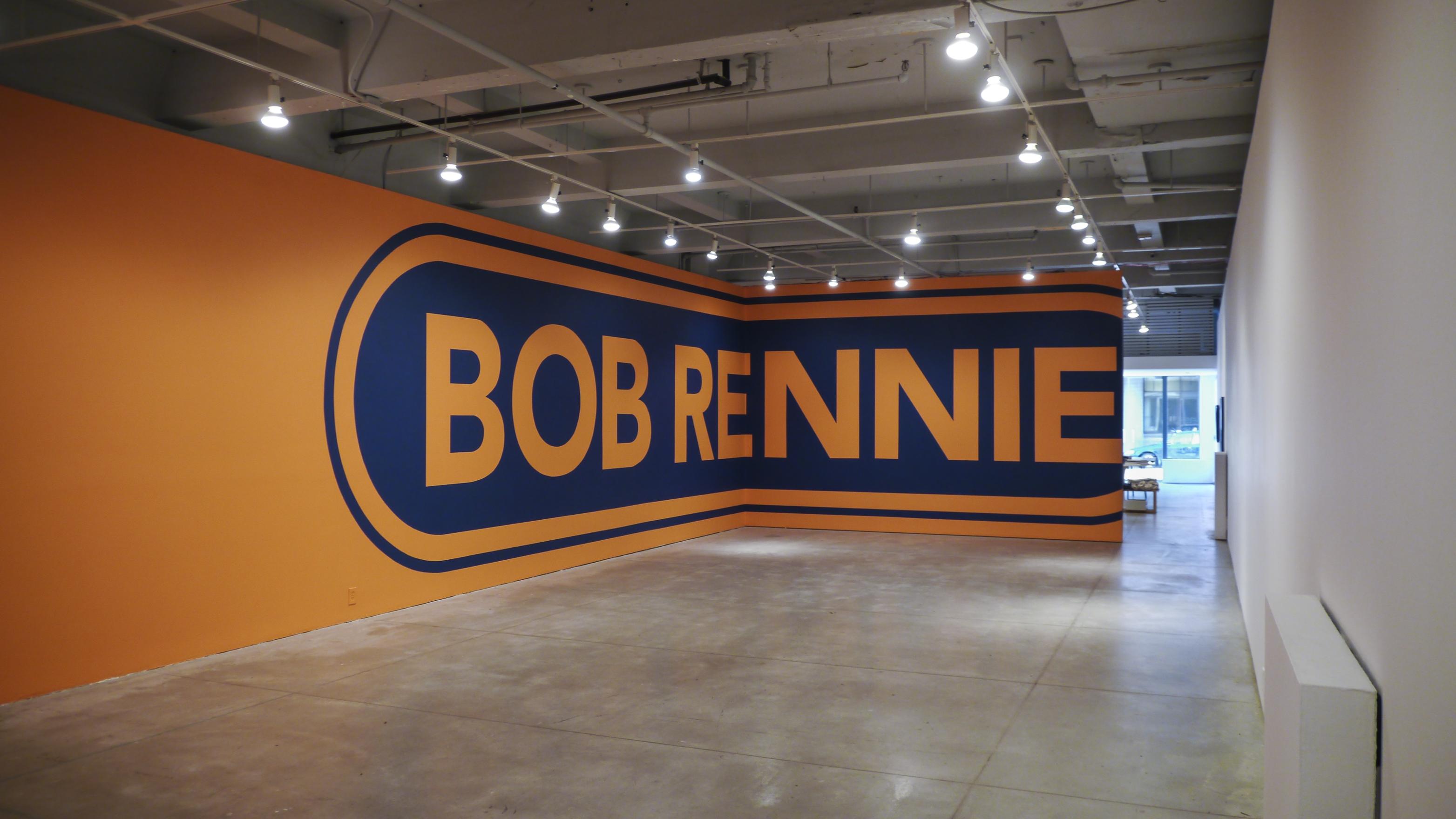 An L shaped gallery wall is painted from floor to ceiling in orange paint. A large oval lozenge shape is painted in navy blue with the name BOB RENNIE stenciled out against the orange background. 