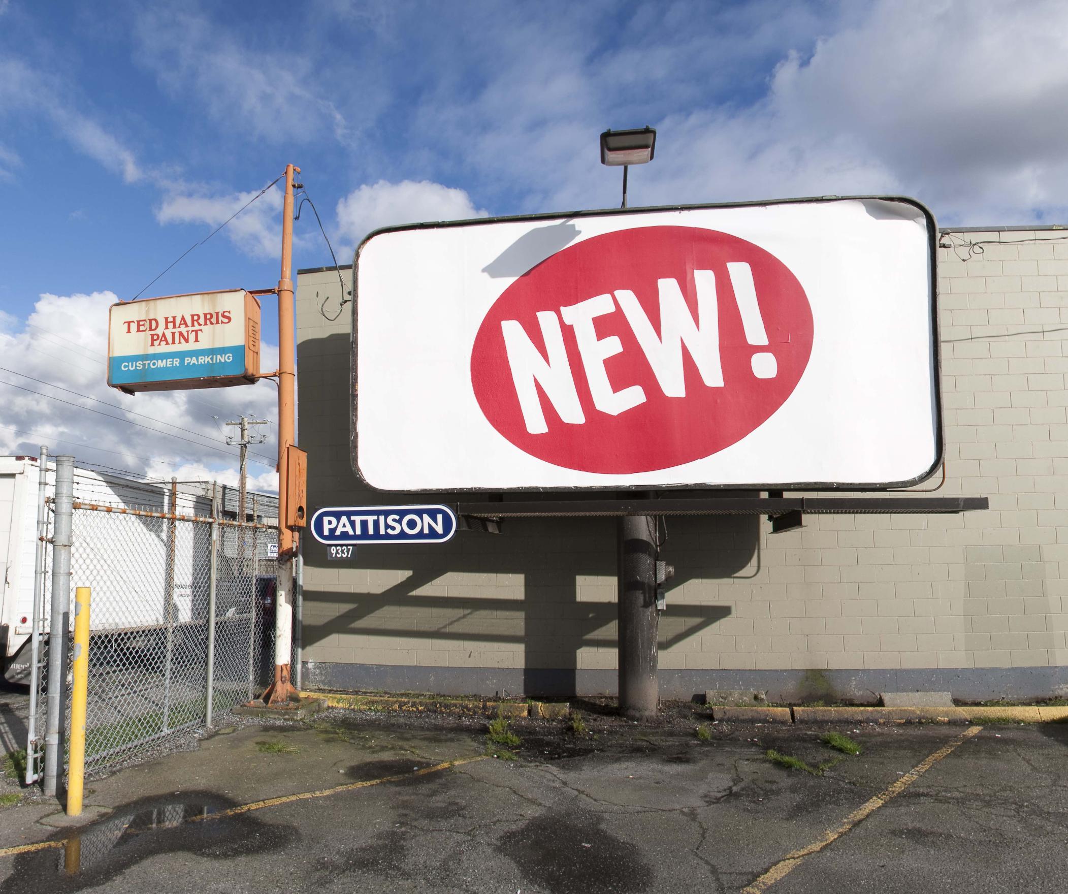  A PATTISON owned billboard stands at the edge of an old parking lot, the word NEW! in sign painters font is written in white in the centre of a large red oval which is in the centre of an otherwise white billboard. This brand-like ovular shape is angled slightly towards the blue sky above. A shiny clean image in a decaying city scape. 