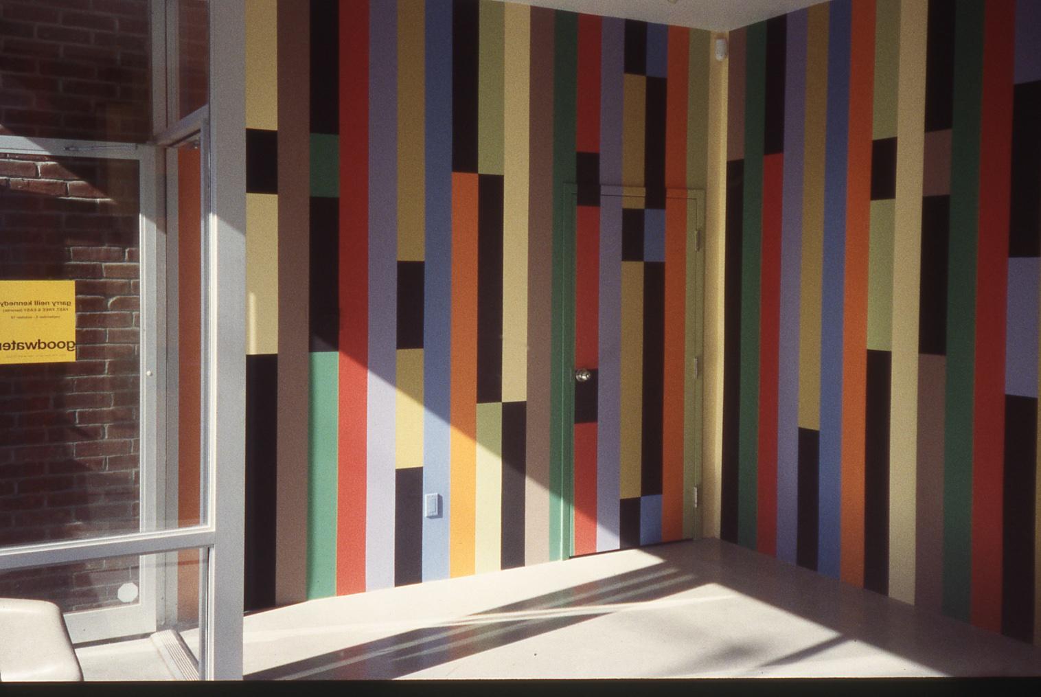 The sun streams in on the walls of a small storefront gallery which are painted from floor to ceiling with the words FAST FREE AND EASY in Superstar Shadow font that has been run through a computer based configuration leaving the letters to condense into thin strips. The colours alternate from light yellows and turquoise, to rust red and orange, and the shadows of each letter are painted in black. 