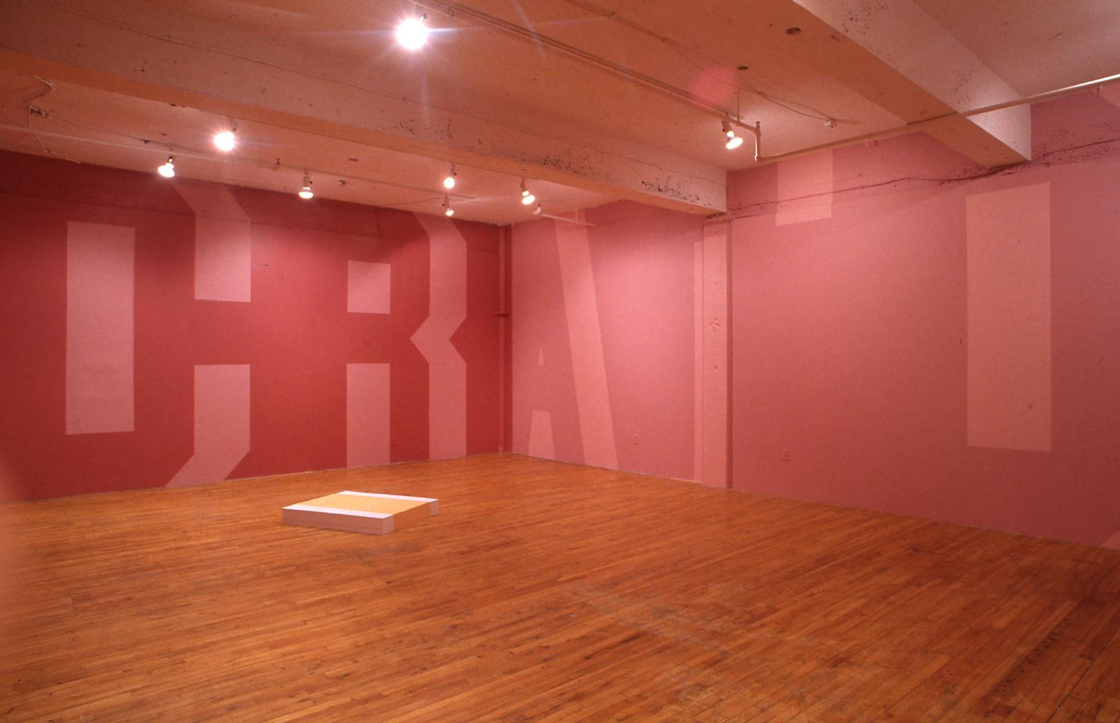 A large wall-text painting envelopes the room in fuchsia and light pink letters which stretch from floor to ceiling, readable letters are C R A T C. A pile of large yellow and white posters are stacked on the hard wood floor of the gallery space. 