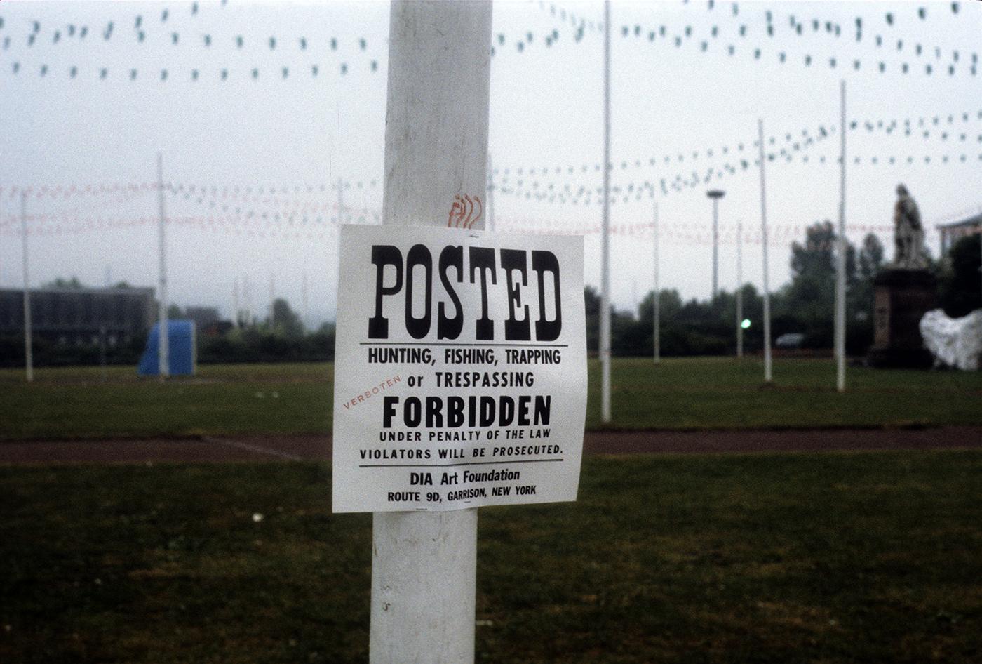 A cream coloured poster has the word POSTED in bold black text at the top, followed by HUNTING, FISHING, TRAPPING, or TRESPASSING FORBIDDEN UNDER PENALTY OF THE LAW. VIOLATORS WILL BE PROSECUTED. DIA Art Foundation, Route 9D, Garrison, New York. The poster is posted on a pole on the grounds of Documenta in Kassel, Germany, with other poles in the background in an empty field. 