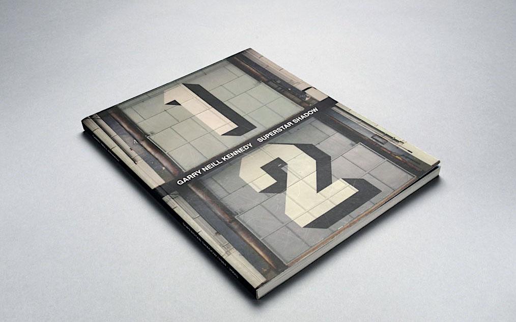  A catalogue lays at an angle on a grey surface. The cover displays two images of wall-text paintings. On top a large number one and below a large number two. Both in Superstar Shadow font painted onto airplane hanger doors in white and black against naval colour grey backgrounds. A black stripe with the book's title separates the two images and reads GARRY NEILL KENNEDY SUPERSTAR SHADOW. 